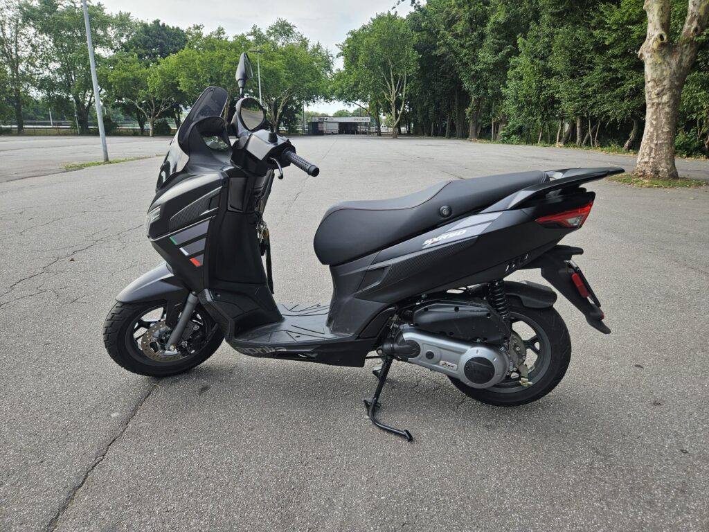 Schwarzer Motorroller auf leerem Parkplatz, umgeben von Bäumen.
