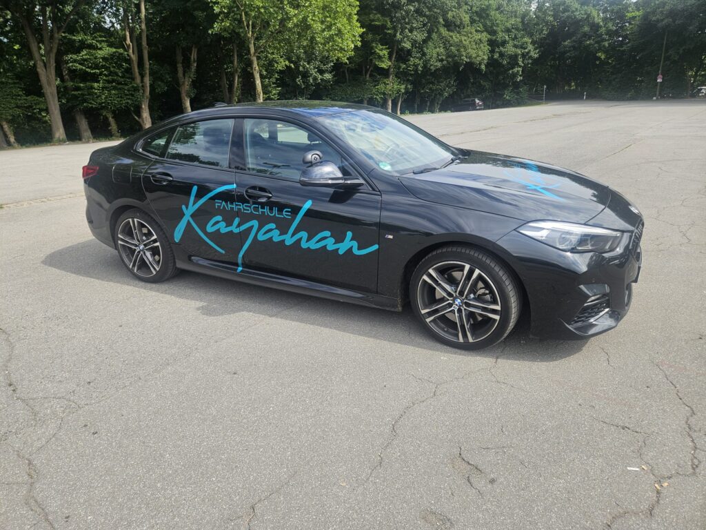 Schwarzes Auto mit blauer Aufschrift Fahrschule, geparkt auf leerem Platz.