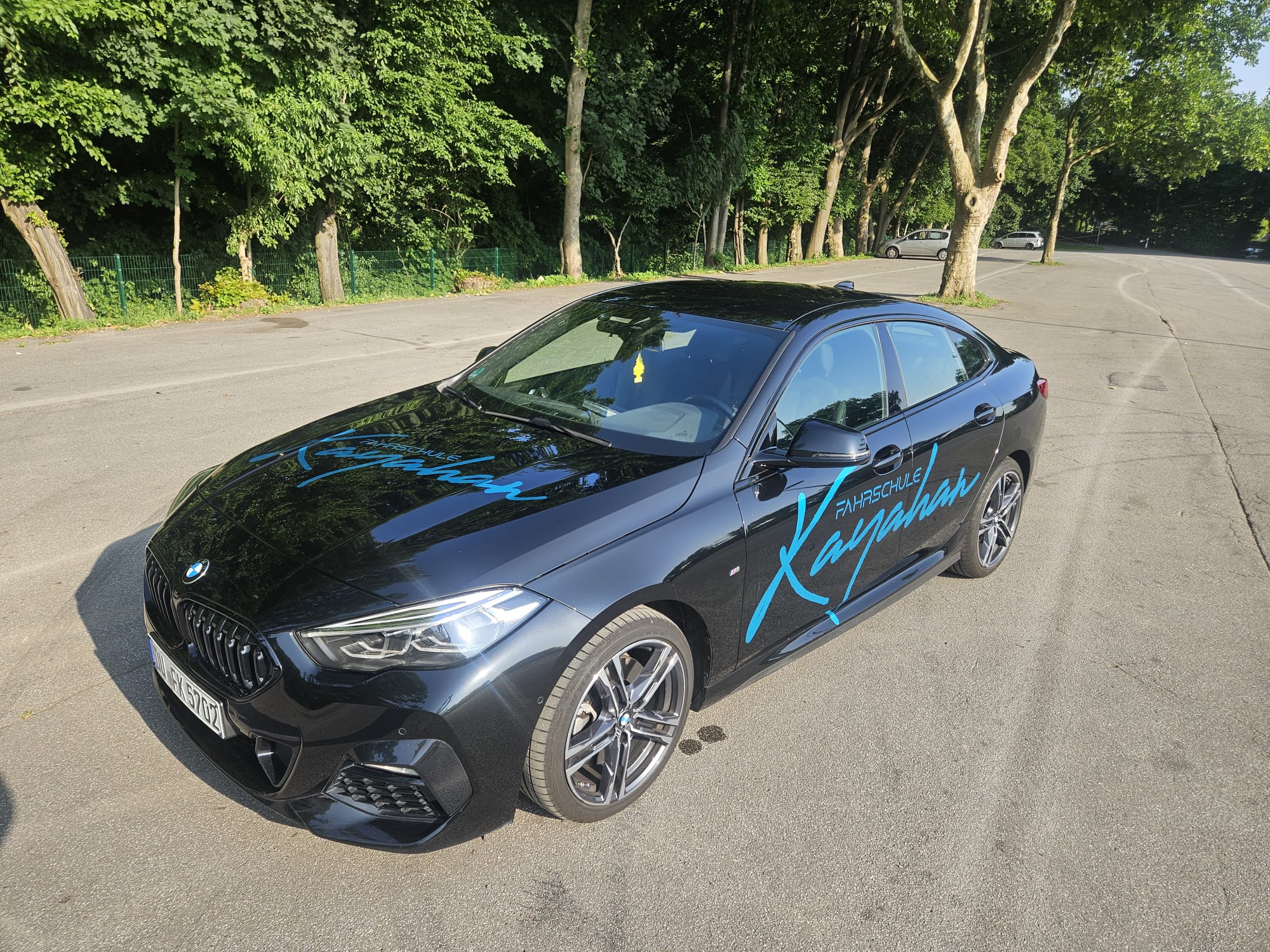Schwarzes Auto, blauer Schriftzug, sonniger Tag, geparkte Straße, Bäume im Hintergrund.