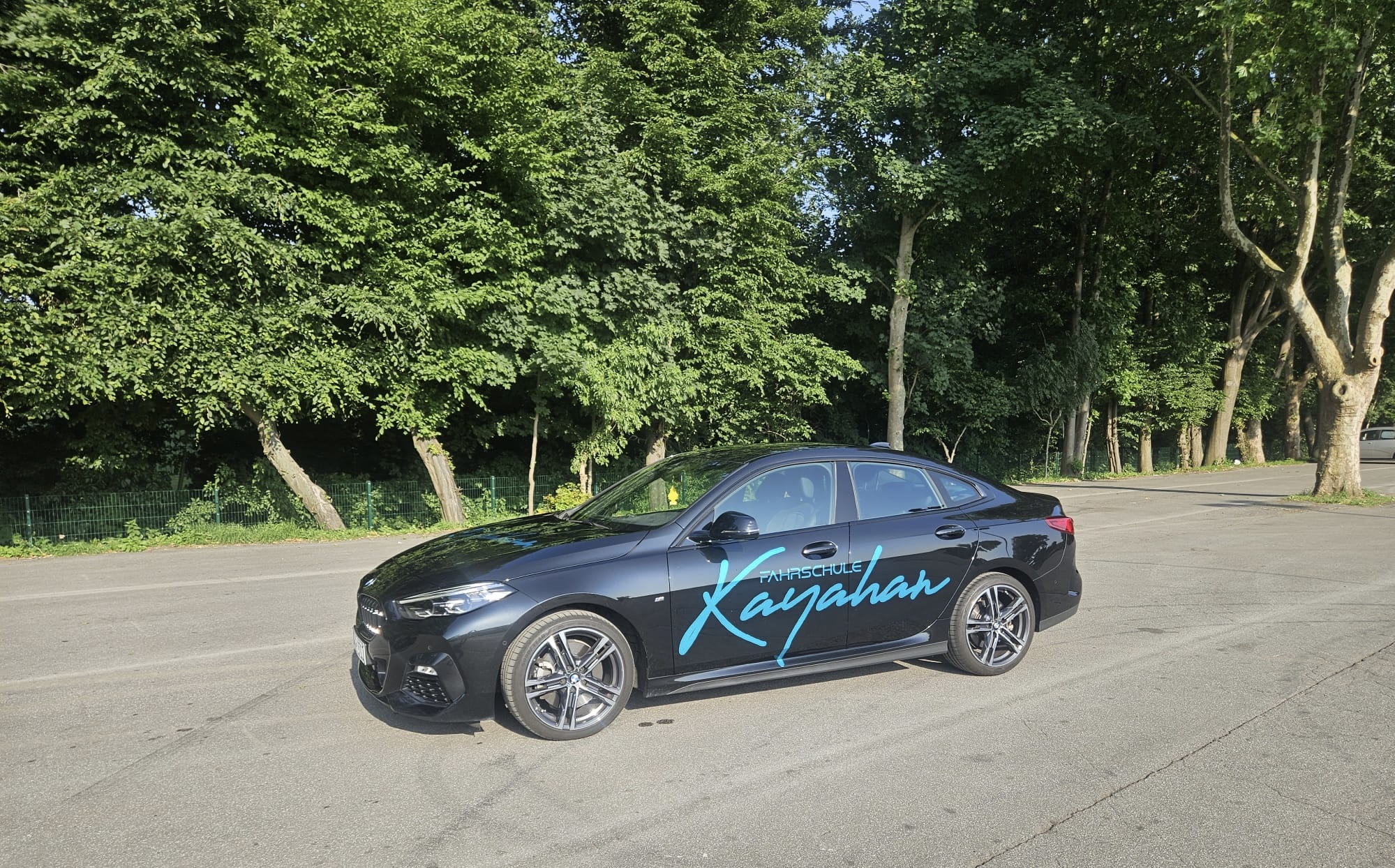 Schwarzes Auto mit Schriftzug, parkend auf Straße, Bäume im Hintergrund, sonniges Wetter.