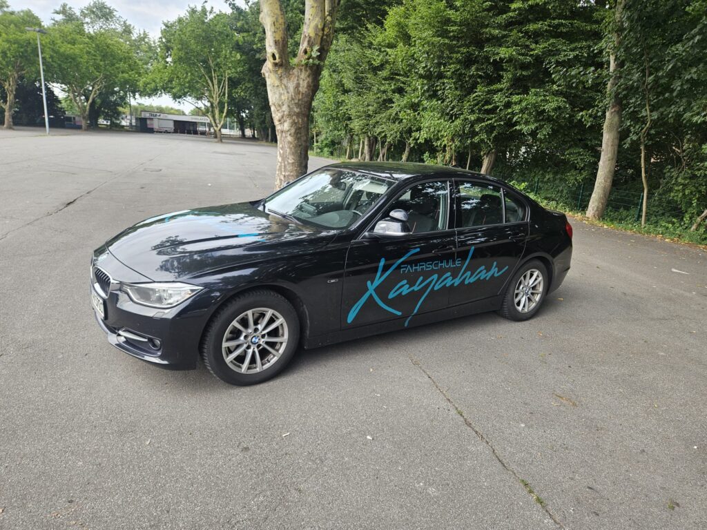 Schwarzes Auto mit Fahrschule-Logo, geparkt auf leerer Asphaltfläche.