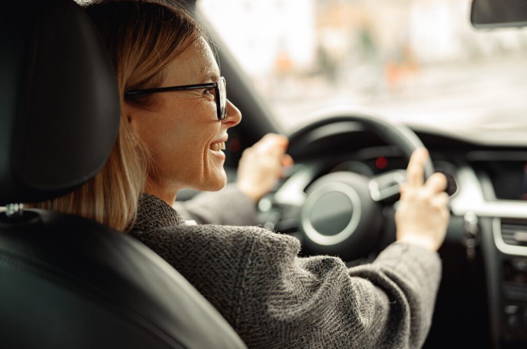 Rückansicht einer lächelnden Frau, die Auto fährt und beide Hände am Lenkrad hält, auf dem Weg zur Arbeit