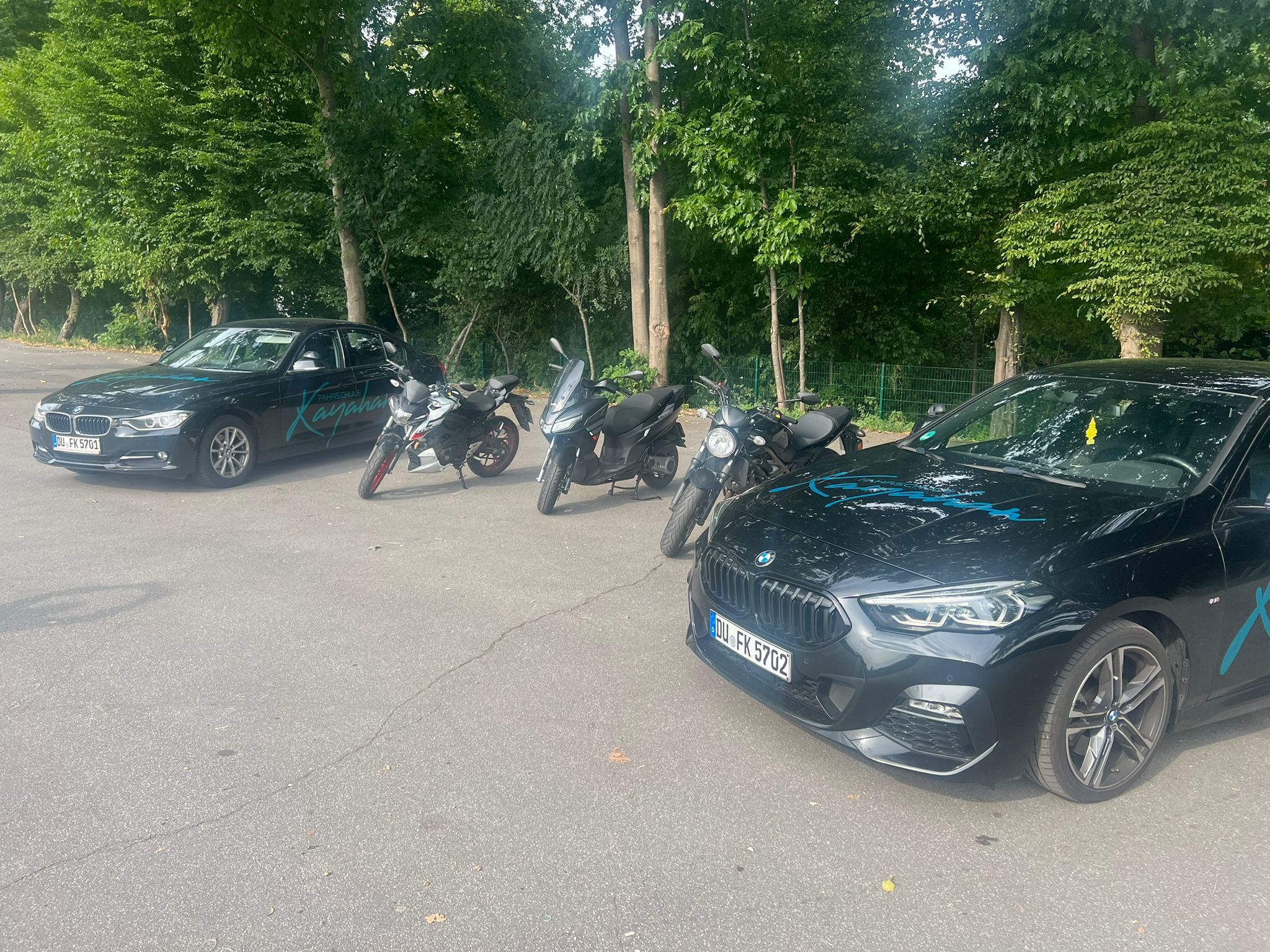 Autos und Motorräder auf Parkplatz vor Bäumen, teils sonnig.